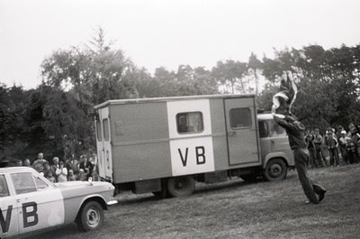 A dynamic 1960s-era photo captures a mid-air jump from a moving truck marked "VB" during a public event. Crowds gather on gra...