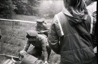 Mid-20th century outdoor scene featuring three men in military-style attire. One kneels assisting a seated individual, while ...