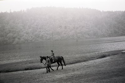 Two riders on horses traverse a misty, rural landscape, likely mid-20th century. The rider in the foreground wears a dark coa...