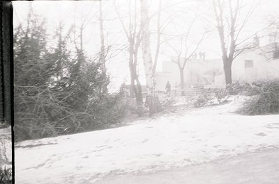 Blurry black-and-white street scene blanketed in snow, featuring leafless trees and a single blurred figure walking. Mid-20th...