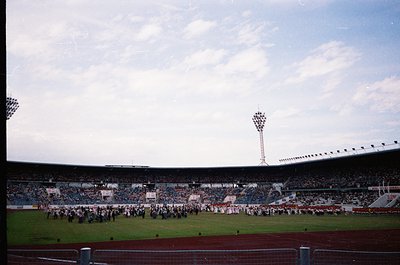 Vintage stadium scene with tiered seating filled with spectators, likely a 1970s European match. Crowds hold flags and banner...