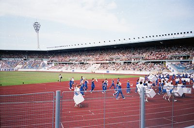 Vintage stadium scene with red artificial turf, featuring a team in blue uniforms celebrating near the sideline. Crowd fills ...