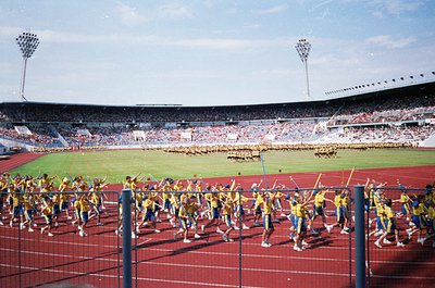 A large-scale synchronized marching band performance in a stadium, featuring uniformed musicians in yellow jackets and blue p...