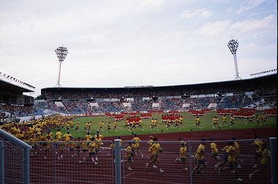 Vintage stadium scene featuring synchronized team drills in yellow jerseys on grass field. Crowd in stands, partially obscure...