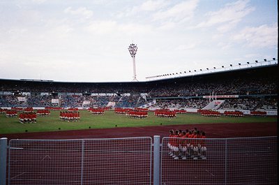 Massive stadium event featuring synchronized marching bands in coordinated red/white uniforms. Crowds fill tiered seating und...