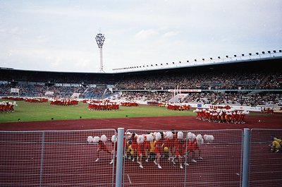 Pre-1990 stadium ceremony featuring synchronized marching bands in red/white uniforms on a red turf field. Crowds fill tiered...