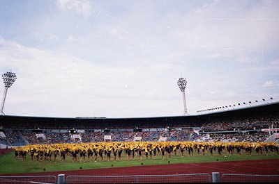 Vintage stadium scene featuring a synchronized dance routine by uniformed performers in yellow and black. Empty stands contra...