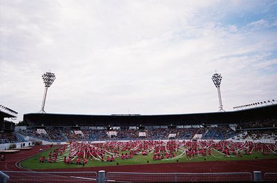 Massive stadium crowd with synchronized dance performance on track, featuring red costumes and choreographed formations. Mid-...