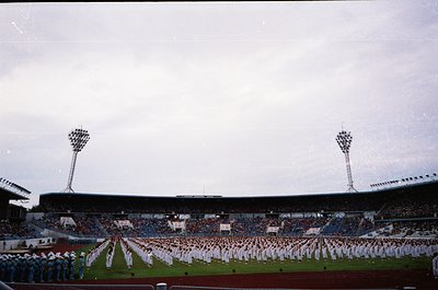 Large stadium filled with uniformed personnel in formation, likely military or ceremonial. Overcast sky with floodlights and ...