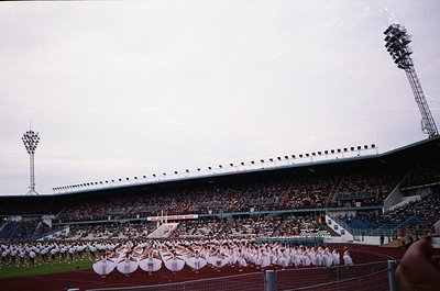 Mid-20th century stadium with tiered seating and floodlights, featuring a large synchronized dance performance by uniformed g...