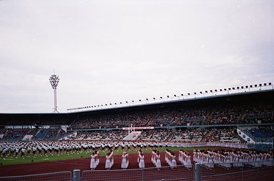 Large stadium filled with spectators, featuring a synchronized dance performance by a group of uniformed women in white on th...