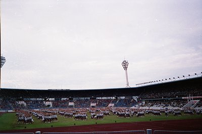 Vintage stadium shot featuring massed marching bands in coordinated uniforms on a grass field. Overcast sky and tiered seatin...