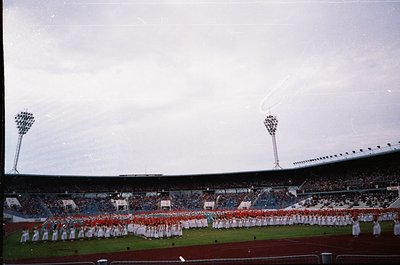 Vintage stadium scene featuring a large marching band in coordinated white and red uniforms performing on a grass field. Over...