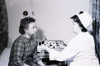 Mid-20th century dental clinic scene: patient seated in chair receiving treatment with early electric dental drill. Nurse in ...