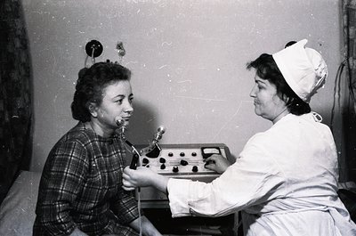 A nurse in a white cap and uniform assists a young patient undergoing dental treatment with a manual dental drill, likely mid...