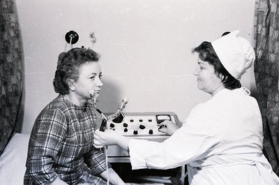 Mid-20th century dental clinic scene: a seated patient receives treatment via a manual dental drill. Nurse in cap and uniform...