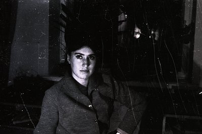 Portrait of a woman in a high-necked, buttoned coat, captured through cracked glass—likely a vintage or urban setting. Her ha...