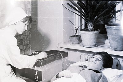 Vintage black-and-white photo of a medical procedure in a clinical setting, likely a 1950s–1960s hospital or clinic. A seated...