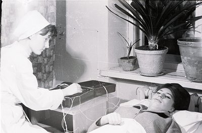 Mid-20th century medical photography: nurse in white uniform adjusts equipment on patient reclining on hospital bed. Medical ...