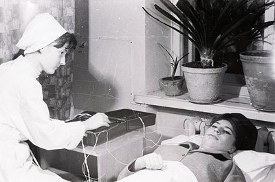Mid-20th century hospital scene: medical professional in white uniform and cap performs electrocardiogram on patient lying in...