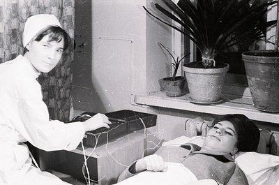 Nurse in 1960s-70s medical attire monitors a patient via electrocardiogram (ECG) in a clinical setting. Indoor hospital room ...
