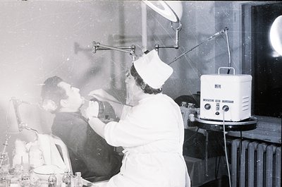 Mid-20th century dental clinic with patient seated in chair, dentist in white uniform applying treatment. Overhead lamp and v...