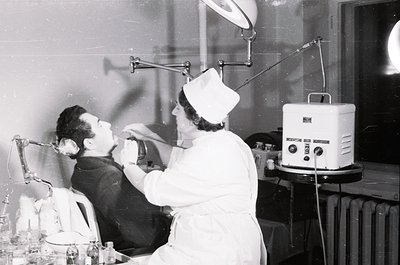 Mid-20th century dental clinic showing patient seated in reclined chair with head tilted back, receiving treatment. Dentist i...