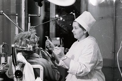 Mid-20th century salon scene: stylist in white cap and coat performs a haircut using vintage clippers on seated client. Indus...