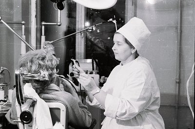 A black-and-white 1960s medical portrait: a stylishly dressed woman in a wheelchair receives a haircut in a salon setting, wh...