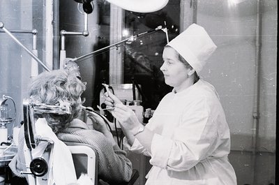 Mid-20th century medical facility: female nurse in cap/gloves assists seated patient undergoing dental treatment with drill. ...