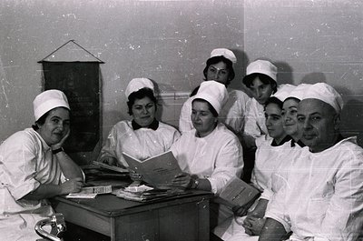 Nursing staff in 1960s-70s uniforms pose indoors, likely a hospital or clinic. White caps, starched gowns, and focused expres...