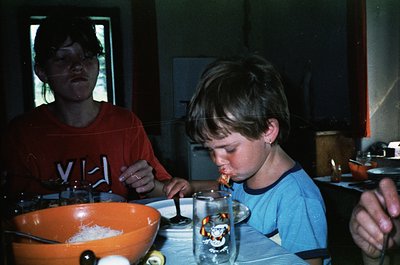 Vintage kitchen scene: two children (early 1980s) focus on a cake with lit candles. Orange bowl, glassware, and retro stove i...