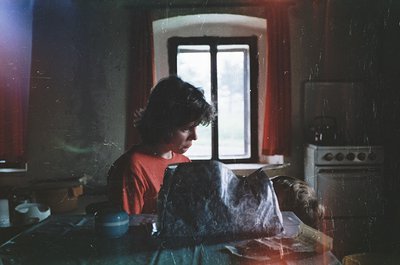 Vintage kitchen scene featuring a woman in a red sweater reading a newspaper while seated at a wooden table. Vintage stove an...