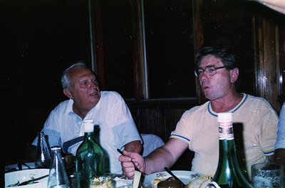 Two men seated indoors at a table with empty wine bottles, likely enjoying a meal or drinks. The man on the left wears a ligh...