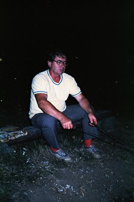 Mid-20th century man in casual outdoor setting, seated on a wooden bench. He wears a white striped polo shirt, dark trousers,...