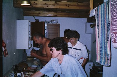 Vintage kitchen scene with three individuals in 1970s-style uniforms, likely lifeguards or resort staff. Wooden ceiling beams...