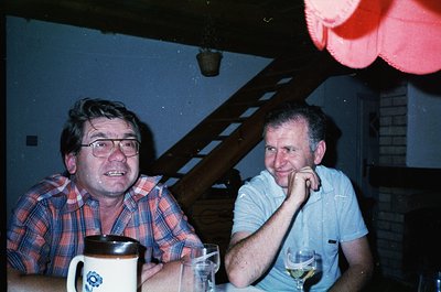 Two men seated indoors, likely in a private home or informal gathering, mid-1970s–1980s. The man on the left wears a plaid sh...