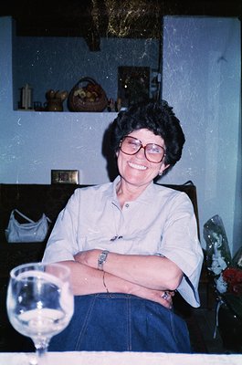 Portrait of a woman in a relaxed indoor setting, likely 1980s–1990s. She wears round-framed glasses, a light button-down shir...