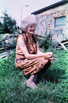Mid-century woman in patterned dress sits cross-legged on grassy lawn, framed by rustic stone wall and garden debris. Likely ...