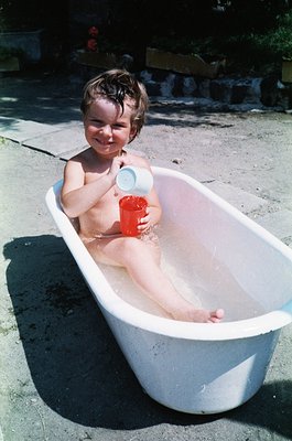 Vintage childhood moment: toddler in outdoor bathtub holding red plastic cup, mid-20th century. Concrete patio with potted pl...