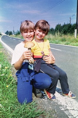 Two children pose on a rural roadside, holding large mushrooms. Boy on left wears a white shirt, blue overalls, and sneakers;...
