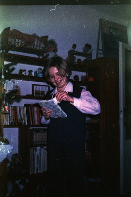 Young girl in 1970s-style school uniform (vest, blouse) holds a book in a dimly lit room with wooden bookshelves filled with ...