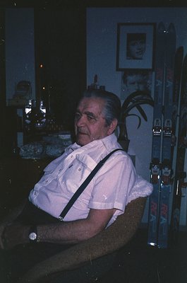 Mid-century man in a relaxed pose, seated in a dimly lit indoor setting. He wears a white button-down shirt with suspenders a...