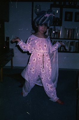 Vintage indoor shot of a child in a pink floral robe, mid-motion, with a headband. Bookshelves and framed artworks in dimly l...