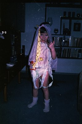 Vintage costume photo of a child in a DIY bride gown, likely 1970s–1980s. White tulle veil, floral sash, and sheer stockings....