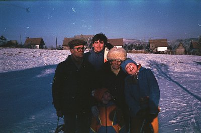 Four individuals pose on a snow-covered ice rink, surrounded by rural buildings. The vintage photo captures 1970s winter atti...