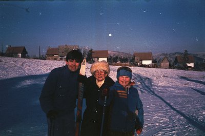 Three individuals pose on a snow-covered slope, likely a ski resort, during dusk. The man wears a dark jacket and cap, the wo...