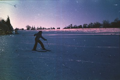 Mid-20th century cross-country skier in motion on groomed trail, silhouetted against twilight sky. Snow-covered forest line i...