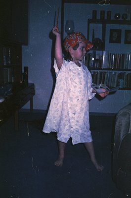 Child in 1970s-style pirate costume (red bandana hat, white floral robe) mid-motion indoors, holding a toy sword. Dimly lit r...