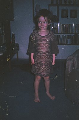Vintage indoor portrait of a young girl in a patterned, knee-length dress, standing barefoot in dim lighting. Bookshelves wit...
